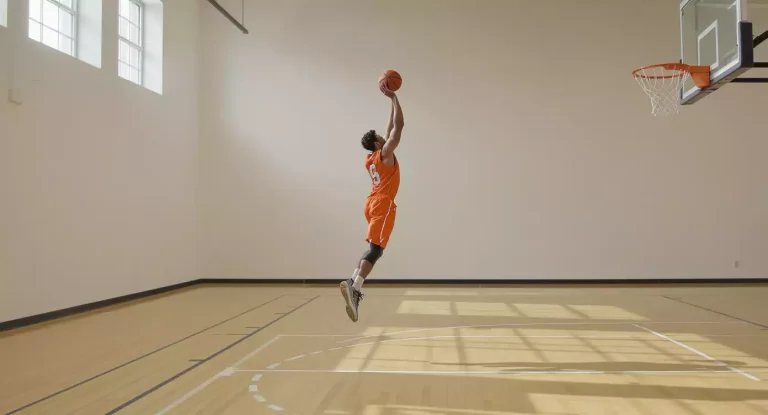 Basketbalspeler in oranje shirt schiet een driepunter in een lege trainingshal met houten vloer