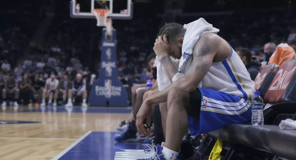 Vermoeide NBA-speler met handdoek op de bench na een wedstrijd, met basketbalveld op de achtergrond