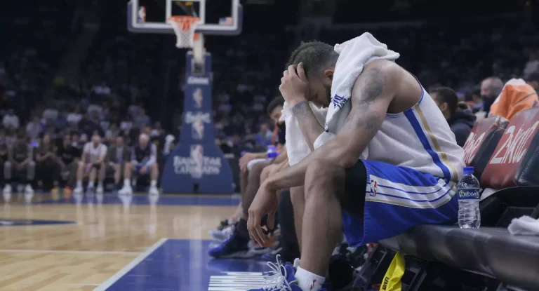 Vermoeide NBA-speler met handdoek op de bench na een wedstrijd, met basketbalveld op de achtergrond
